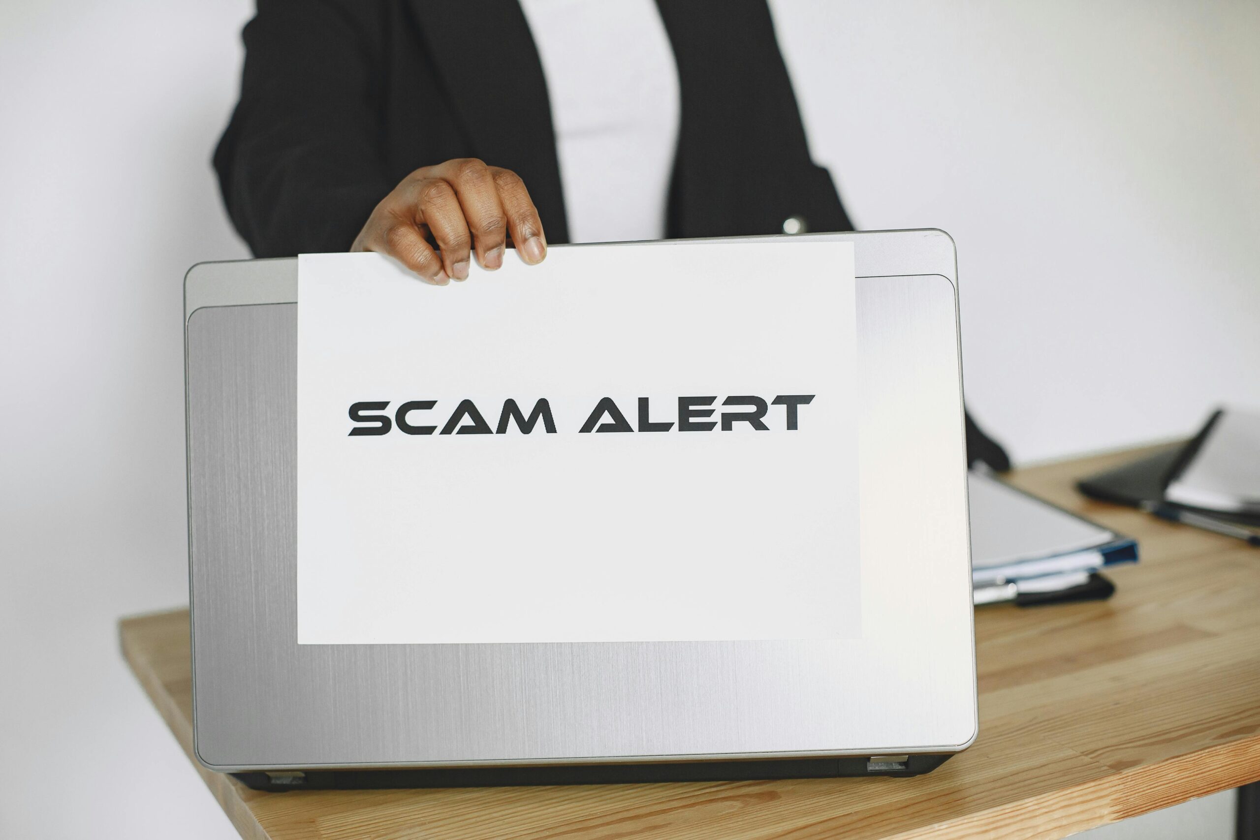 A person holding a sheet of paper that reads “Scam Alert” in front of a laptop, symbolizing a warning about online scams and cybersecurity awareness in the workplace.