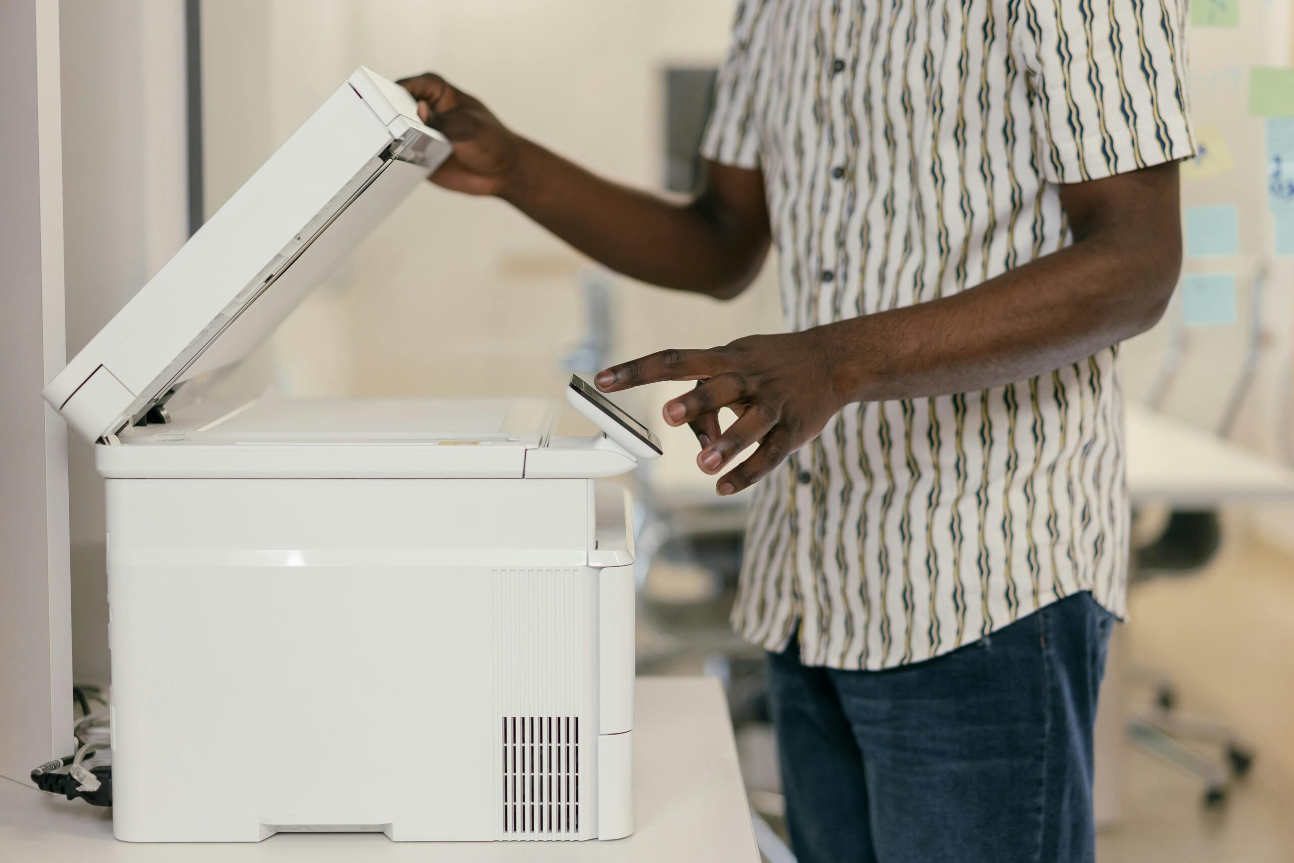 A person standing beside an office printer, lifting the lid and using the control panel, representing troubleshooting or printing in a workplace setting.