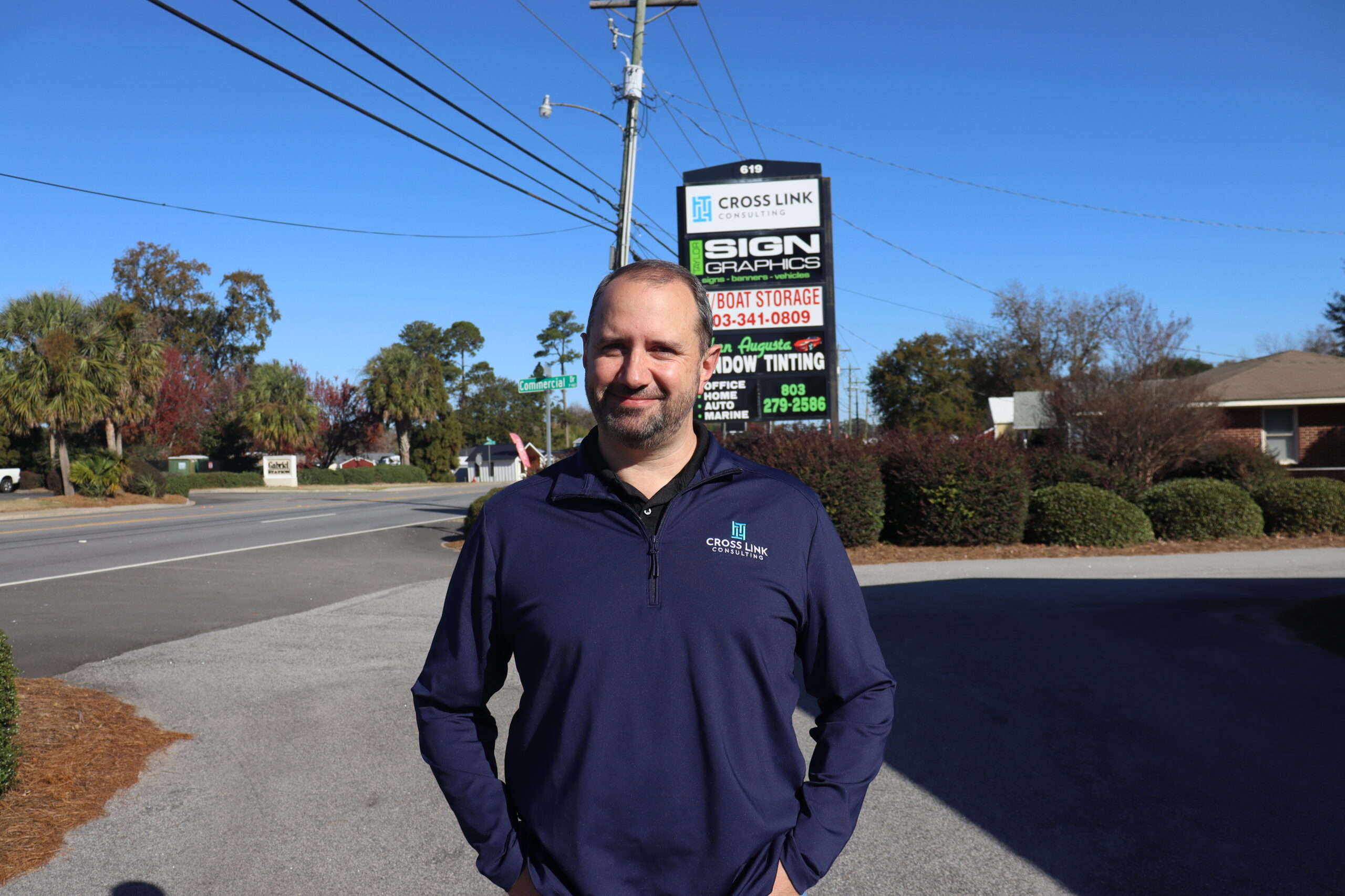 Local IT professional wearing a Cross Link Consulting jacket standing outdoors near the company sign, representing a locally owned and operated managed service provider serving the Augusta and North Augusta community.